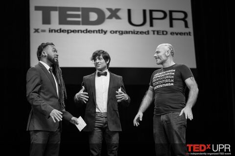 Carli on stage hosting TEDxUPR, in a bowtie, with two speakers beside him